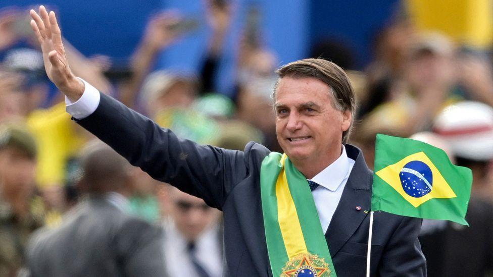 Žair Bolsonaro sa predsedničkom lentom preko sakoa i zastavom Brazila u jednoj ruci, maše pristalicama
