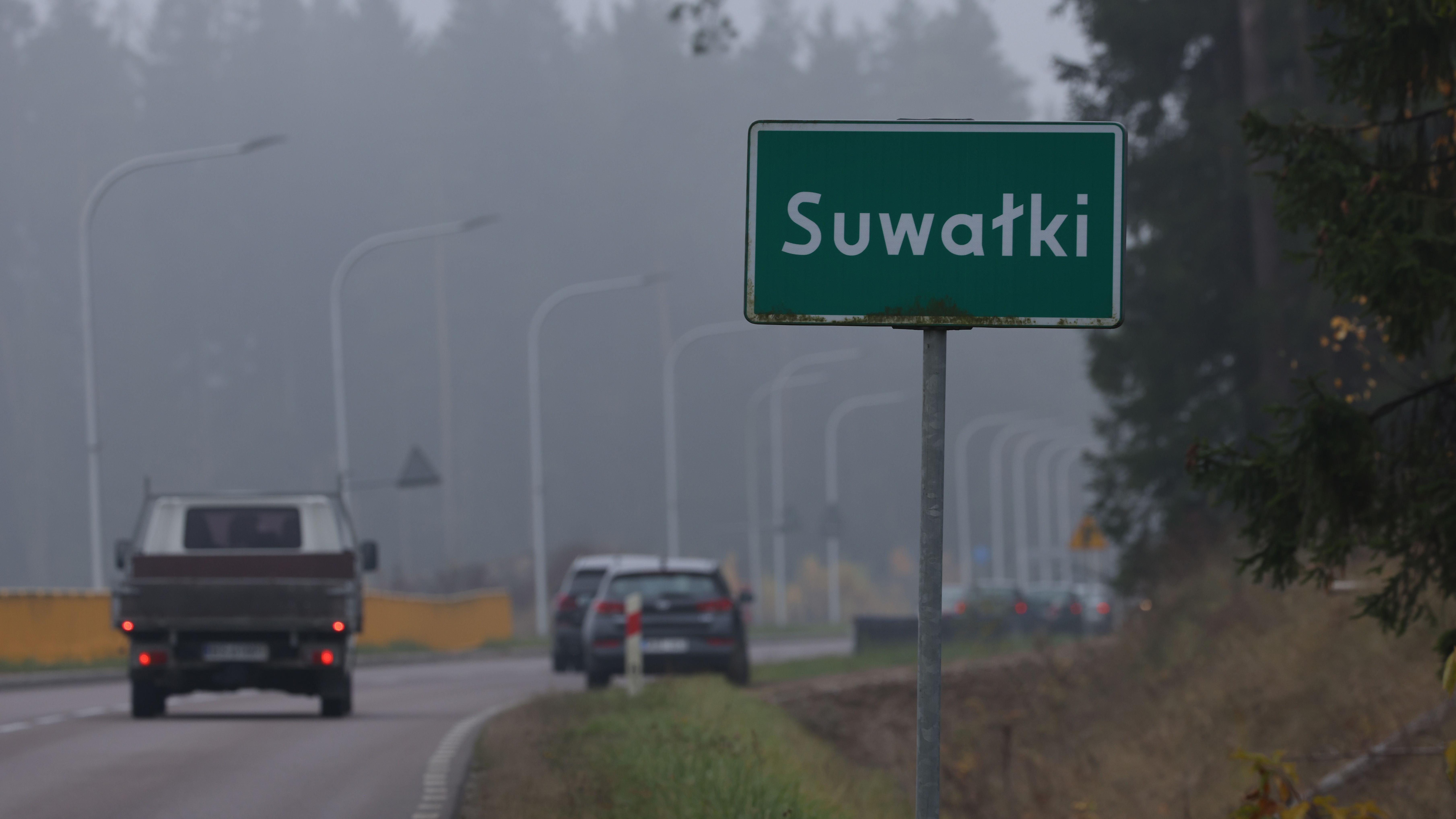'Zona opasnosti' u Evropi za koju malo ljudi zna: Suvalkski koridor 4 Dva automobila prolaze pored grada na putu sa zelenim saobraćajnim znakom za Suvalki u Poljskoj. U pozadini se nalaze drveće i ulična rasveta
