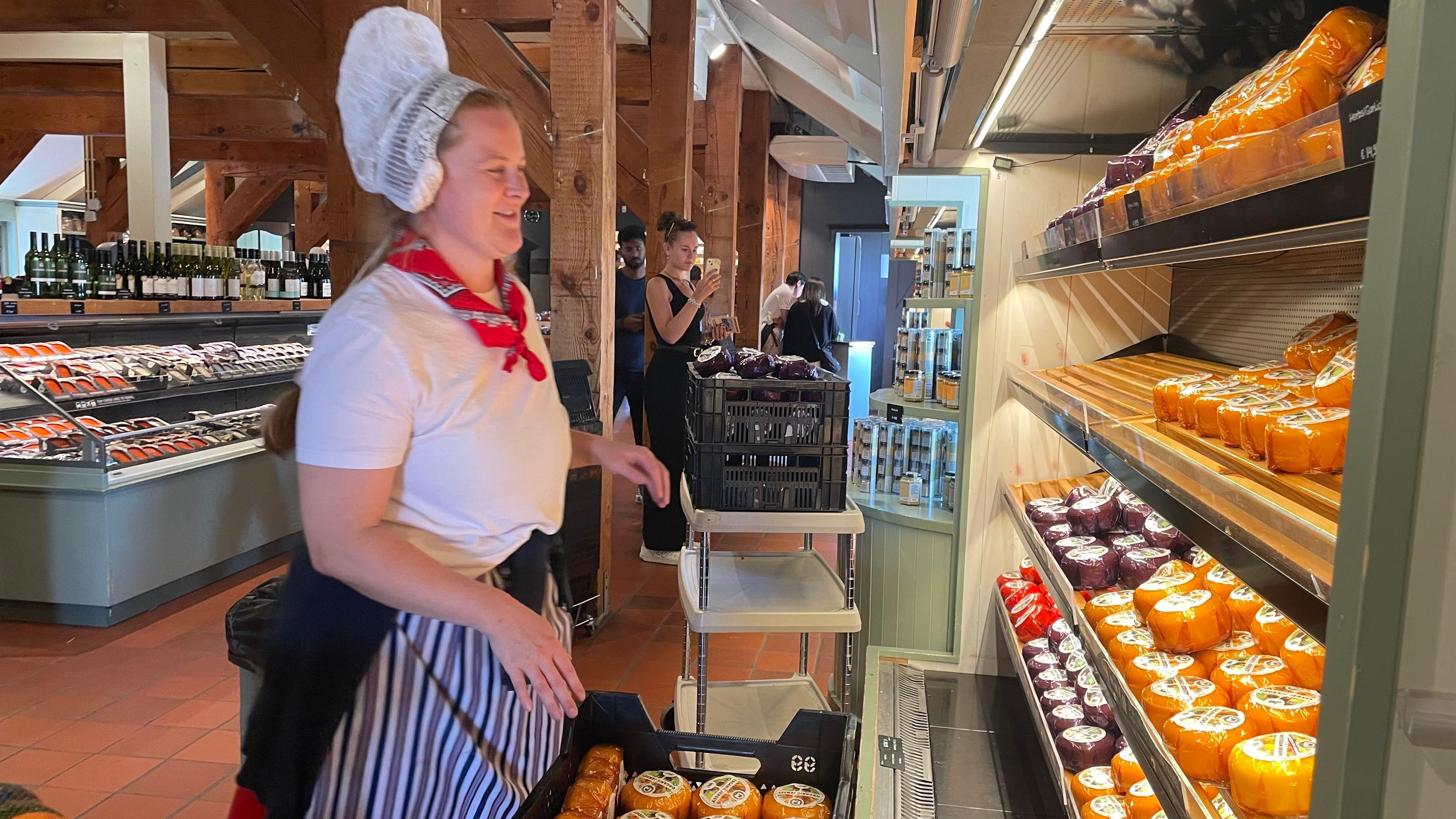 Da li biste platili 17,5 evra da gledate vetrenjače 3 žena obučena u tradicionalnu nošnju - bela majica i bela kapica, sa crvenom maramom oko vrata ispred rafova na kojima su poređani sirevi