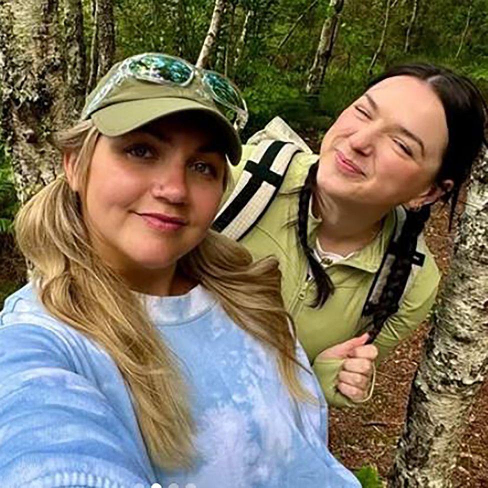 San, vežbanje, hidriranje - da li zaista moramo da se držimo preporučenih dnevnih doza 4 Emili i Lusi - dve žene stoje, okružene drvećem. Jedna nosi svetloplavu majicu i ima dugu plavu kosu. Druga je obučena u zelenu majicu i nosi ranac.