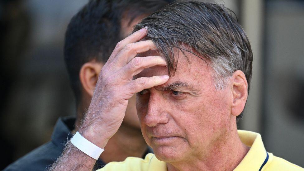 Bolsonaro u krupnom kadru rukama prolazi koroz kosu, nosi žutu majicu i gleda u zemlju