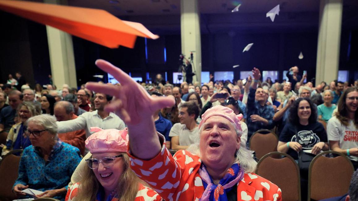 Čovek iz publike u crvenom sakou sa belim srcima baca papirni avion