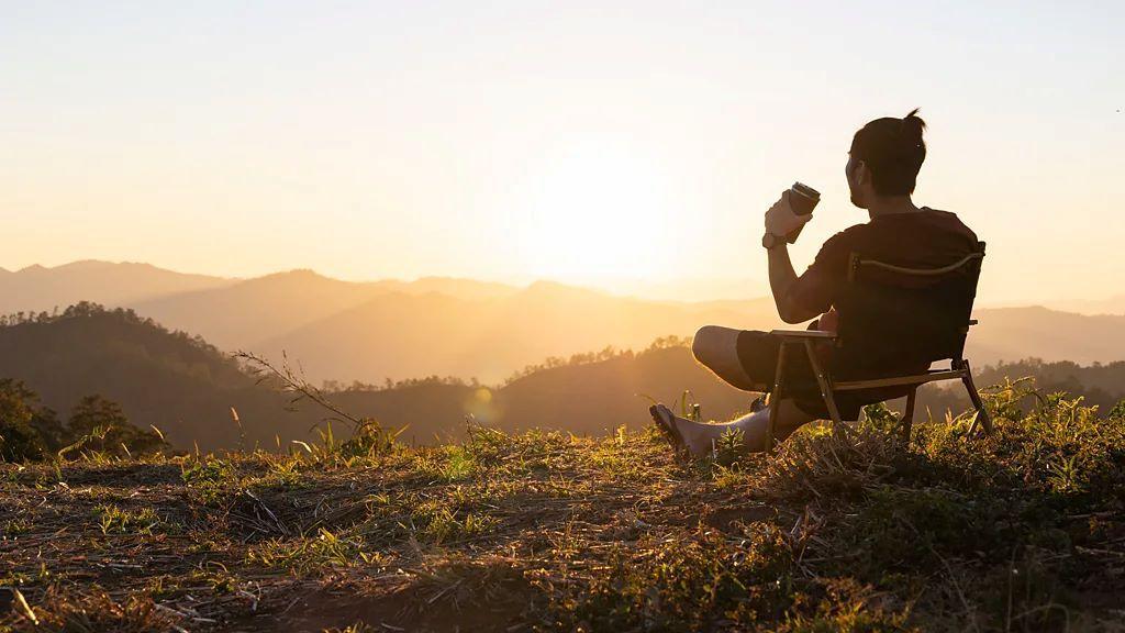 Manje kafe, živopisniji snovi 1 muškarac sedi na stolici na uzvišenju okrenut kameri, u ruci drži kafu dok gleda u sunce koje izlazi