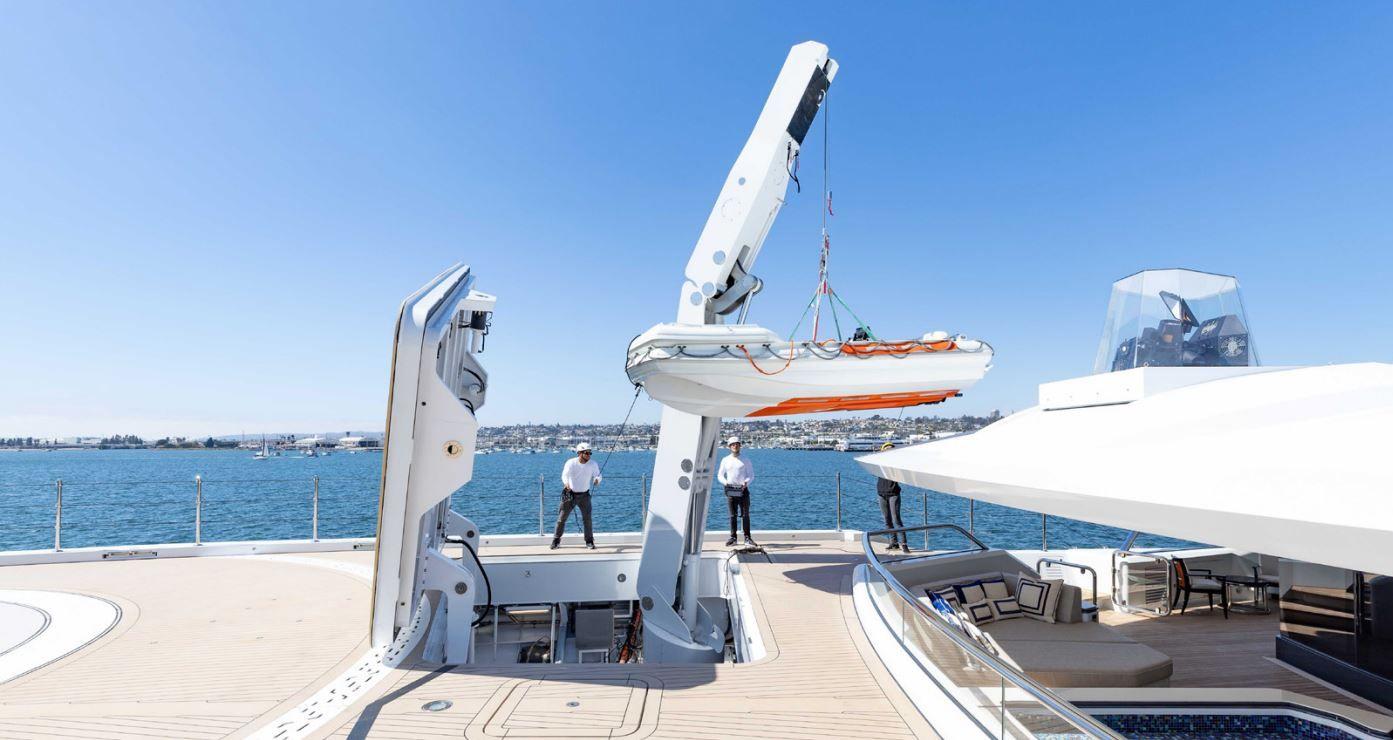 'Beskonačni' bazen, helidrom, osam soba: 'Superjahta ruskog milijardera' na aukciji 12 Dva muškarca u mornarskim uniformama stoje na palubi, sa čamcem podignutim iznad palube. Ivica helidroma je u levom delu fotografije, a desno je prostor za sedenje sa belim pokrivačem. Sunčan je dan, vidi se kopno u daljini.