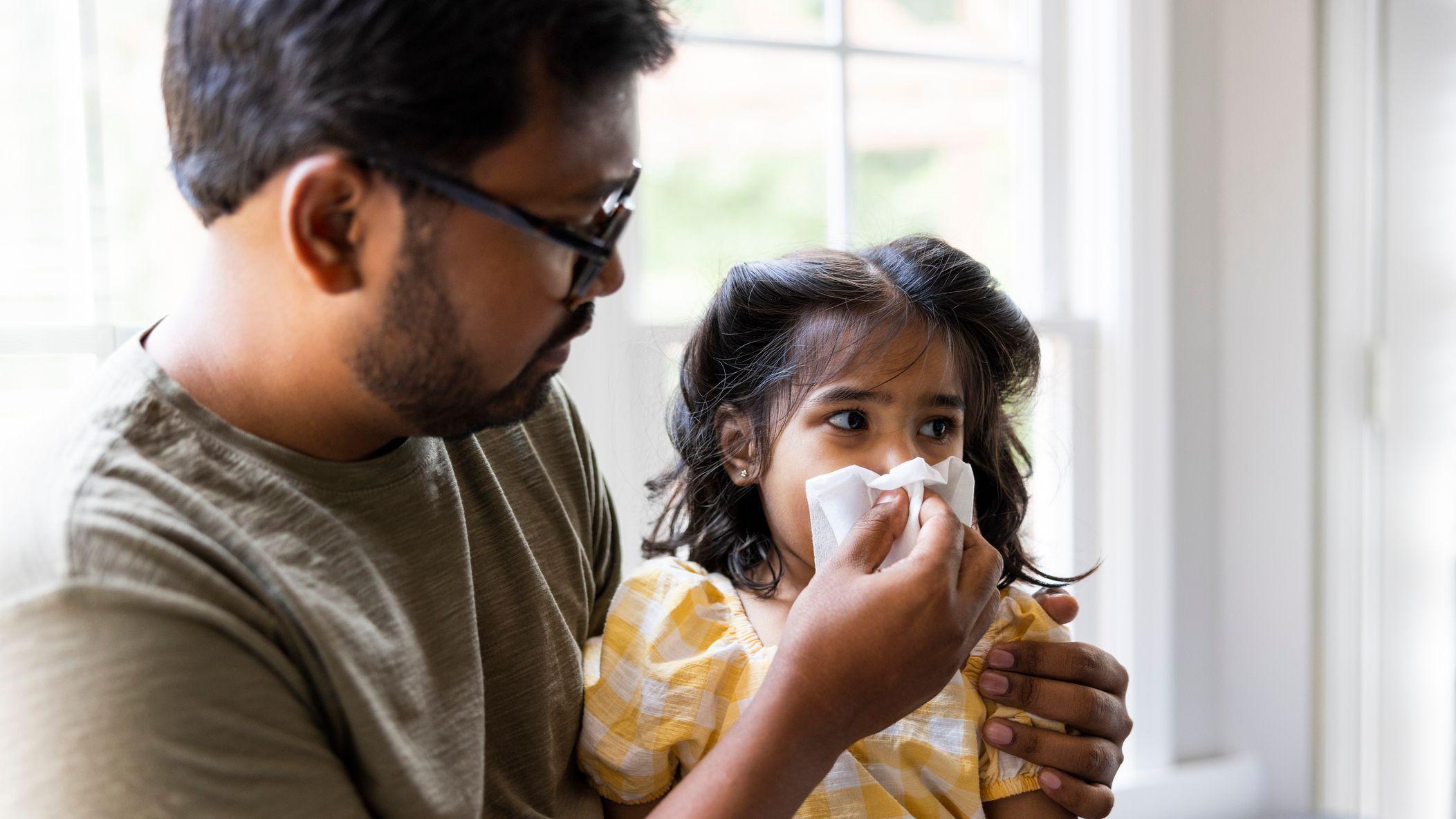 Otac drži ćerku u krilu dok joj pomaže da ispuca nos maramicom. Ona nosi žutu haljinu, a njen tata crne naočare i krem majicu.