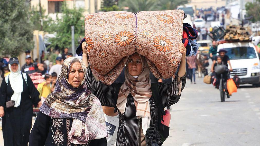 Raseljenja u Gazi 'neuporediva sa bilo čim decenijama unazad' 5 Dve žene sa maramama na glavi, jedna od njih nosi smotani dušek na glavi, u daljini natoverana kola stvarima