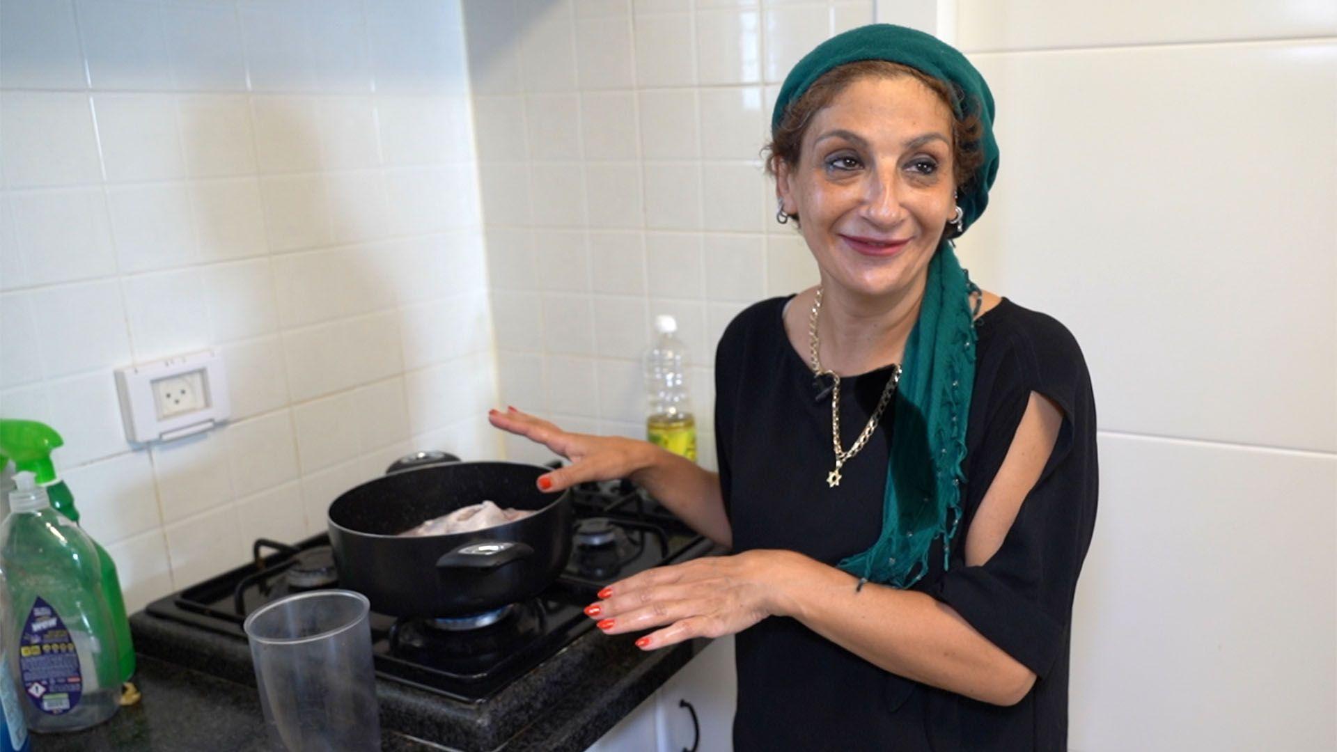 Žena stoji pored otvorenog crnog lonca na šporetu. Nosi zelenu maramu oko glave, crnu majicu i zlatnu ogrlicu sa zvezdom. Iza nje je boca ulja, a zidovi su prekriveni belim pločicama