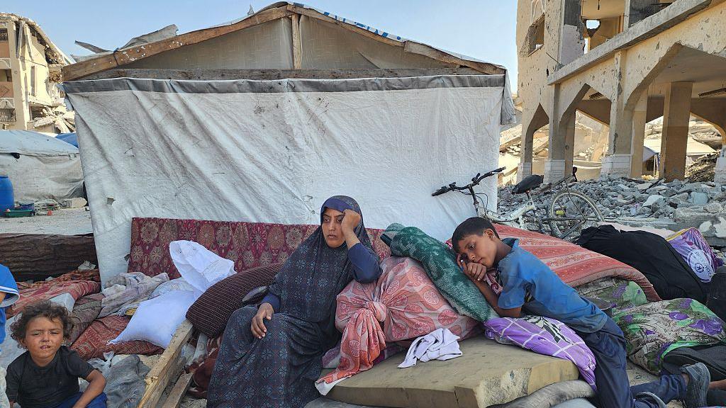 Raseljenja u Gazi 'neuporediva sa bilo čim decenijama unazad' 6 Žena sedi između dvoje dece, jednog predškolskog uzrasta i starijeg dečaka. Oni se smeštaju na gomilu dušeka i stvari ispred improvizovanog skloništa napravljenog od platna i komadića trave. Stariji dečak se oslanja na jastuk kao da pokušava da zaspi, žena naslanja glavu na ruku sa tužnim, umornim izrazom lica. Ostaci uništenih zgrada mogu se videti sa obe strane skloništa. Snimljeno u oblasti al-Mavasi, 17. septembra 2025.