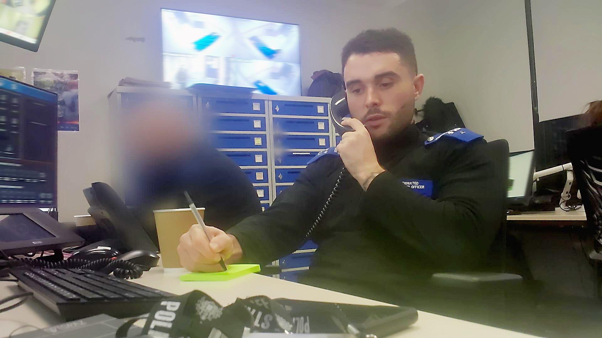 Rory Bibb, seen working at his desk while wearing the uniform of a designated detention officer, a navy jumper with epaulettes in royal blue. He is talking on the phone in front of a computer, making a note on a yellow Post-It note. Behind him is a colleague whose face is blurred