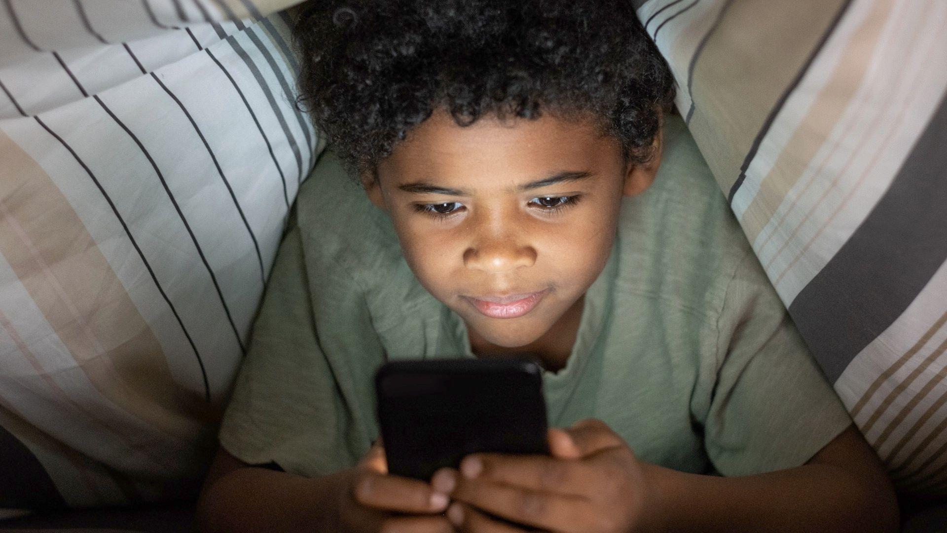 Crnoputi dečak, kovrdžave kose, pod jorganom je i gleda u pametni telefon