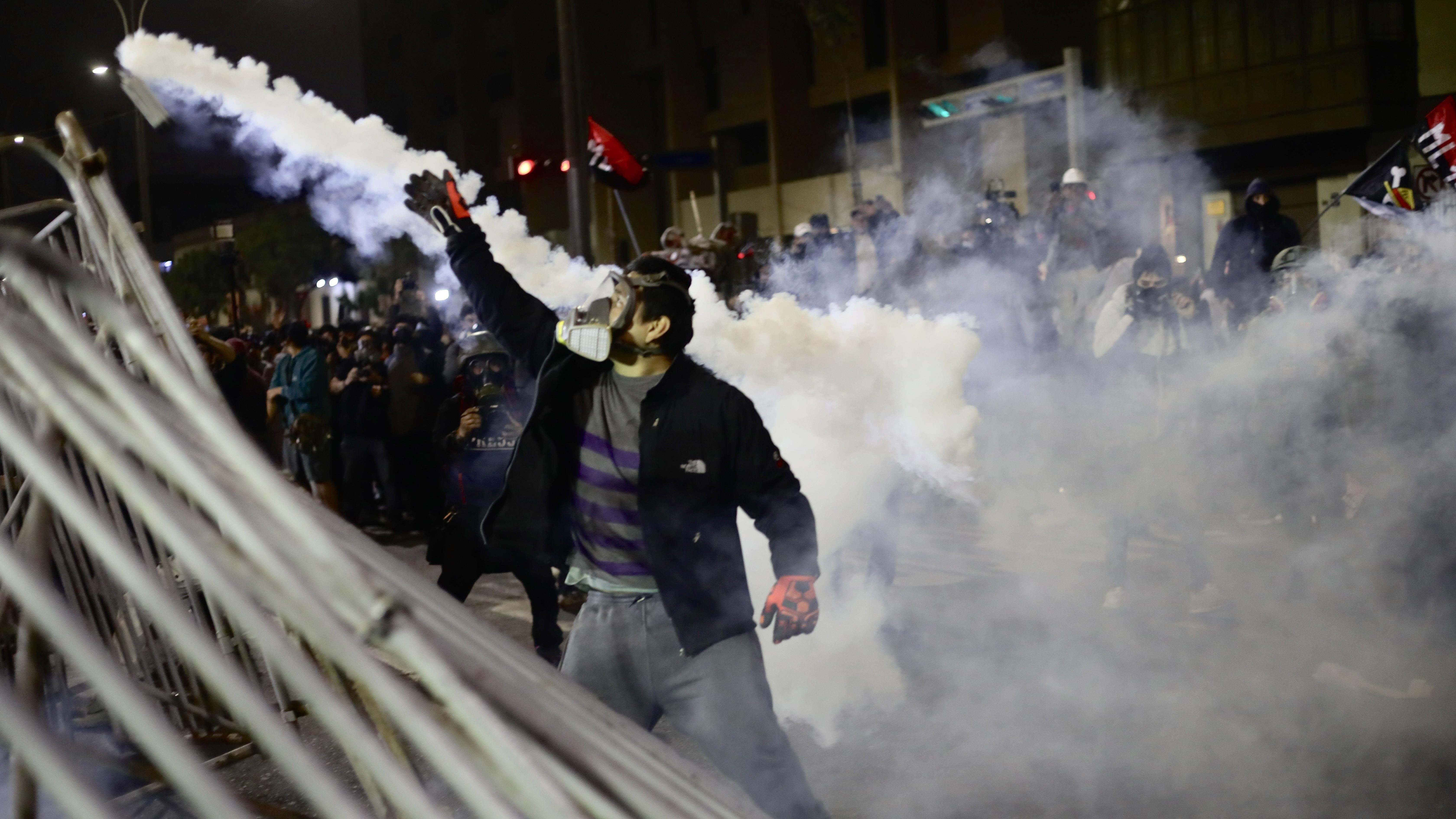 Demonstrant u Peruu sa maskom i rukavicama podiže desnu ruku dok stoji u suzavcupored delimično srušene metalne ograde. Grupa ljudi je okupljena iza njega