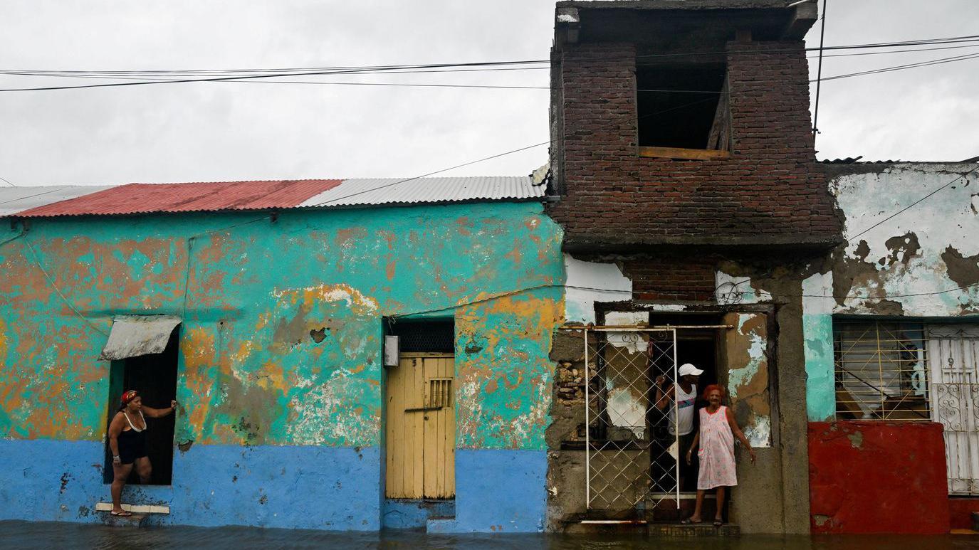 Troje ljudi stoji na dovratku uronule jednospratne kuće, oko njih je svuda voda