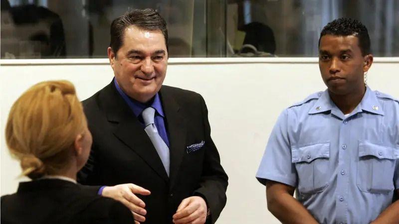 Ko je bio Nebojša Pavković, general osuđen za ratne zločine na Kosovu 1 Nebojša Pavković u haškoj sudnici u crnom odelu i plavoj košulji, smeje se i pruža ruku. Pored njega je policajac u uniformi, stoji sklopljenih ruku. Ispred Pavkovića je žena, u crnoj garderobi, okrenuta leđima, sa narandžastom kosom