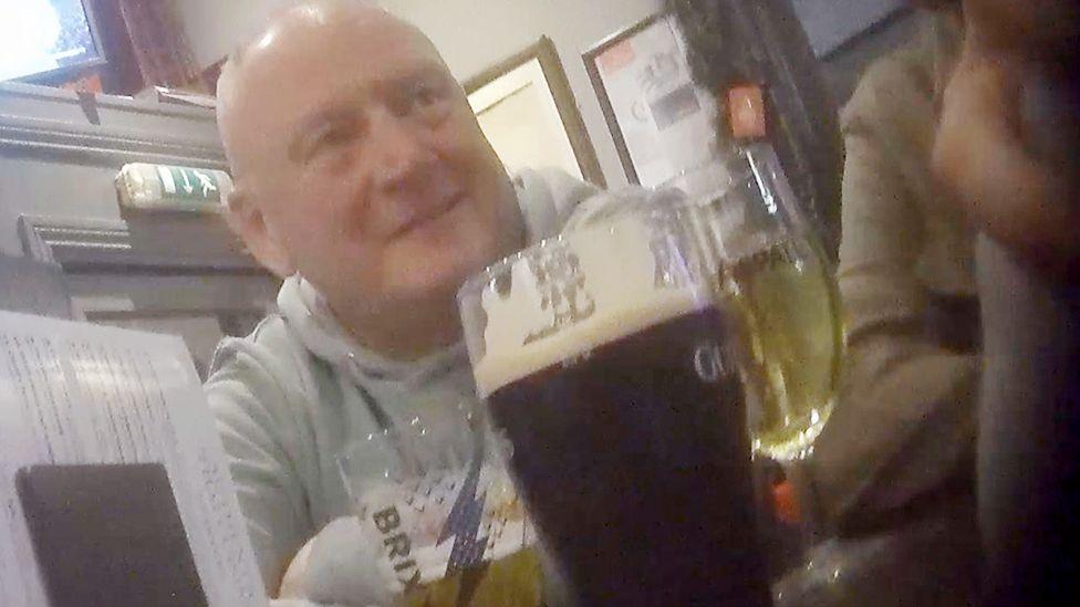 A hidden camera shot of PC Brian Sharkey, a white man wearing a light grey hooded top, sitting at a pub table with multiple glasses and menus in front of him and other colleagues just visible out of shot