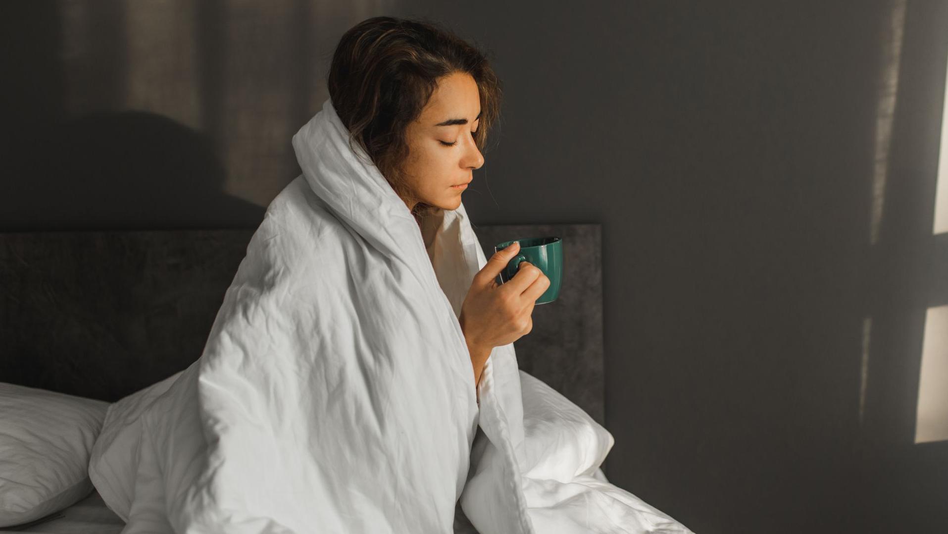 grip, žena ima grip, devojka ima grup, žena sedi pored kreveta umotana u beli jorgan, dok drži zelenu šolju. Ima tamnu kosu i pogled joj je spušten ka podu