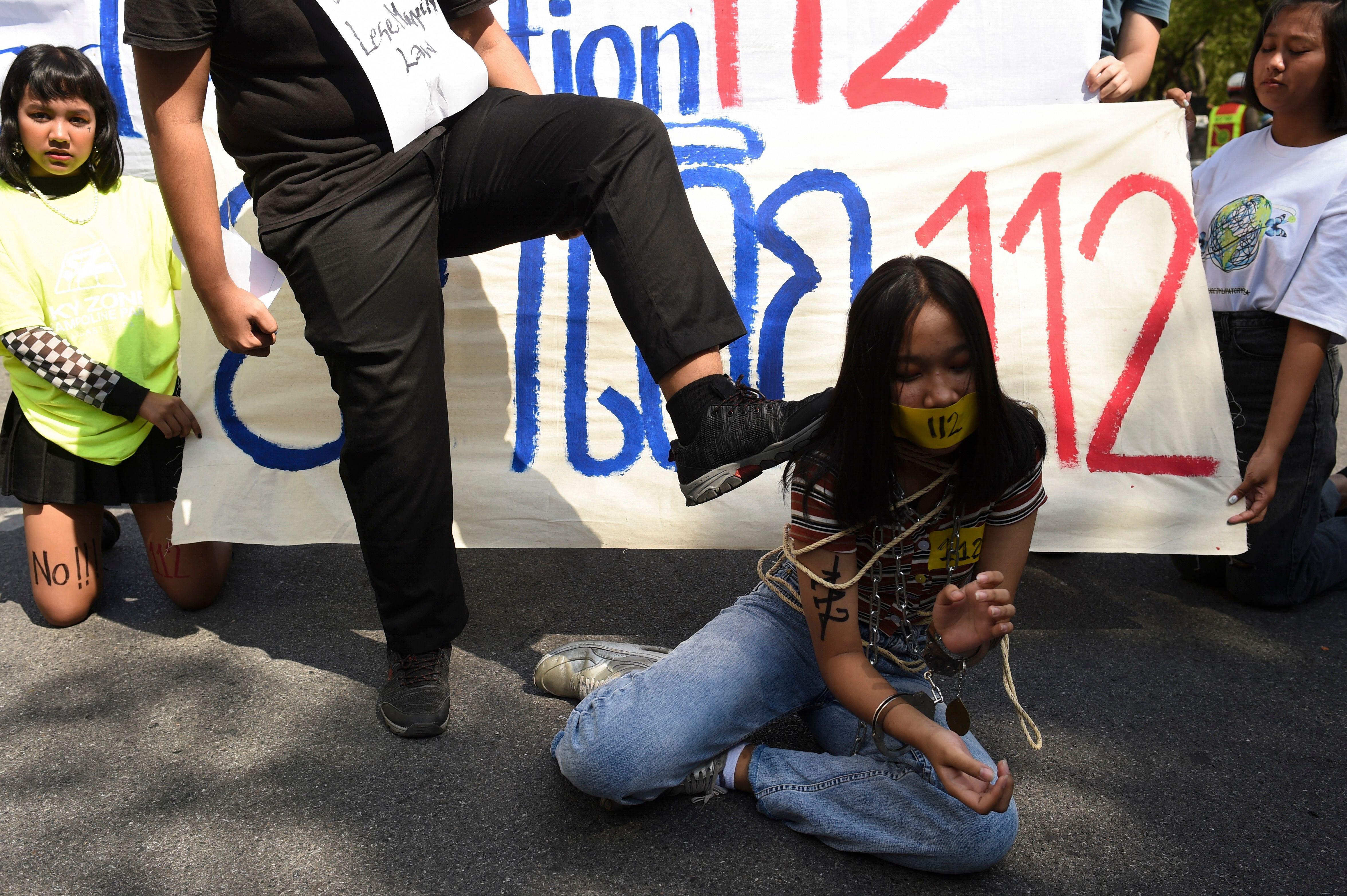 Demonstrant stavlja nogu na rame mlade demonstrantkinje koja kleči na zemlji vezana konopcem. Na transpatrentu iza nje je broj 112, član tajlandskog zakona koji kriminalizuje vređanje monarha