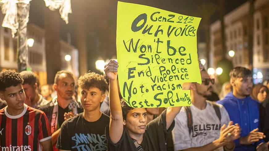 Mladi ljudi na protestu u Rabatu, u Maroku, 3. oktobra 2025. godine, na demonstracijama na kojima su tražili reforme u obrazovanju i zdravstvu i bune se protiv korupcije. Jedan demonstrant drži transparent na kojem piše: „Gen Z 212 - Naš glas neće biti ućutkan. Kraj policijskog nasilja. Ne rasizmu. Da solidarnosti.“