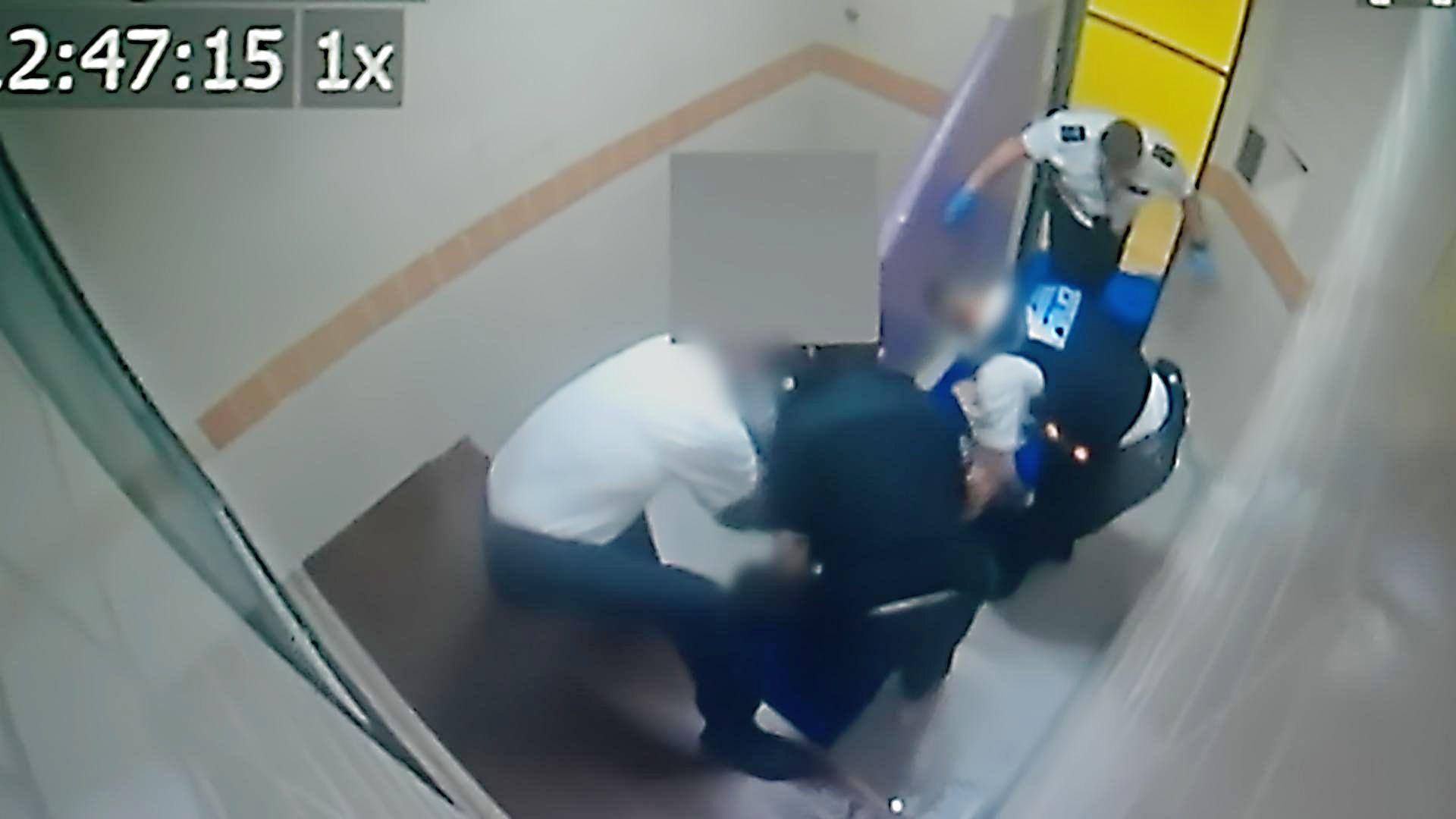 A still of a CCTV image showing three officers in a small cell, who appear to be restraining a suspect, barely visible in the crowd. At the top right of the image, by the open door of the cell, is a man identified as Sgt Steve Stamp, who can be seen stomping down twice