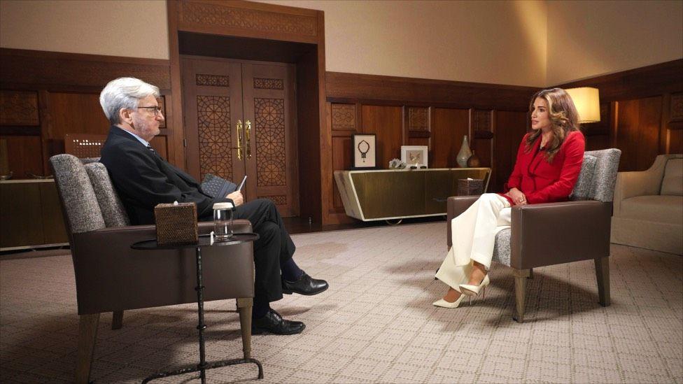A man with greying hair in a black suit sits to the left interviewing a woman with long brown hair, a red shirt and smart white trousers and shoes