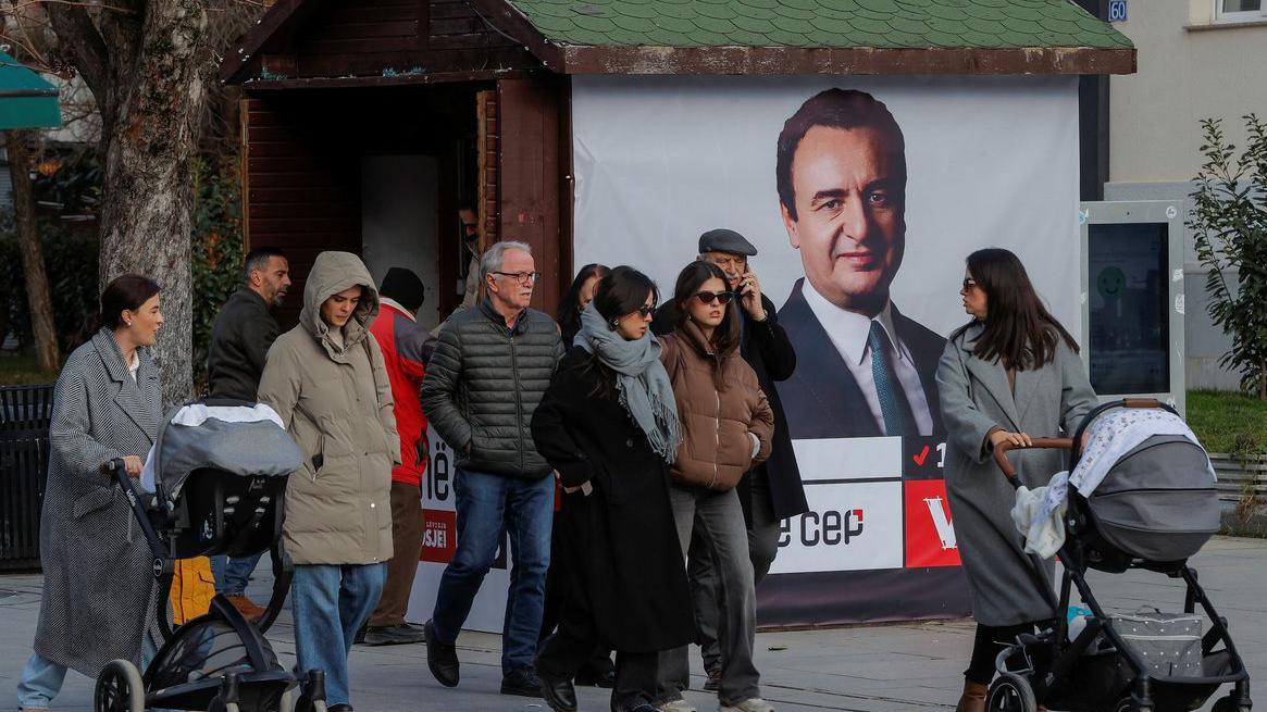 Lokalni izbori na Kosovu: Ključne stvari koje treba da znate 1 Dve žene guraju kolica, između njih je nekoliko devojaka i dva muškarca, u pozadini bilbord sa slikom Aljbina Kurtija