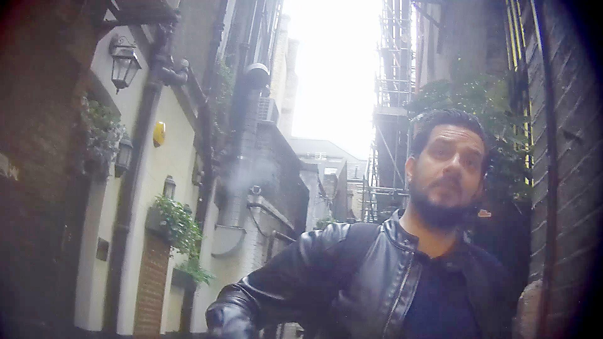 A hidden camera shot taken at chest height looking slightly upwards at PC Martin Borg, a white man with dark hair and a short, dark beard, standing in what appears to be an alleyway outside a pub, wearing a dark T-shirt and a black leather jacket