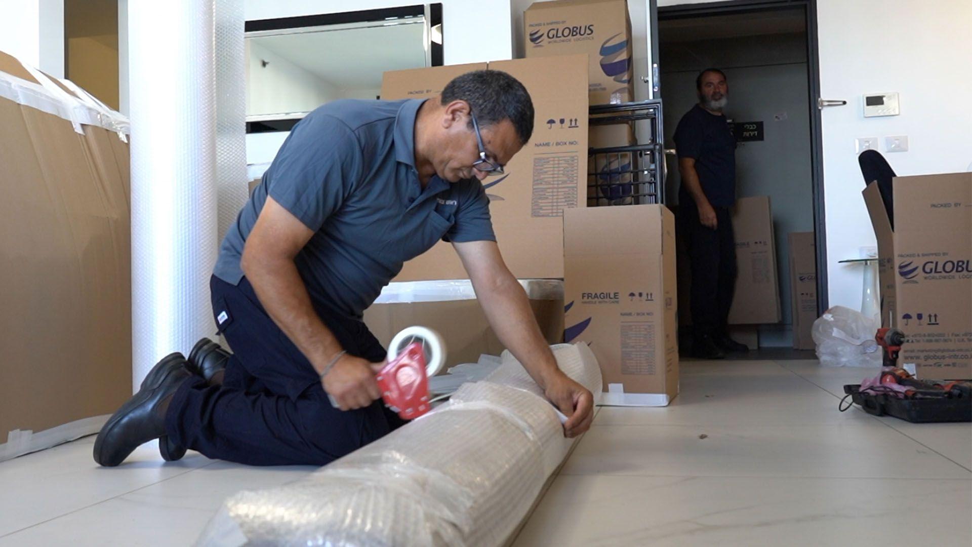 Čovek u tamnim pantalonama kleči na podu i lepi selotejpom veliki predmet umotan u mehuričastu foliju. Iza njega su kartonske kutije i još jedan čovek koji posmatra