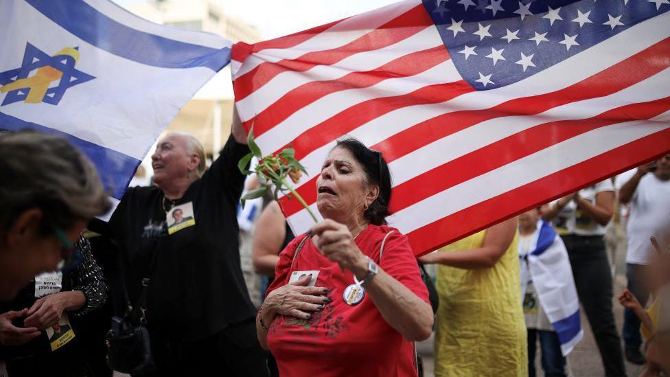 Kako je Tramp postigao dogovor za Gazu, koji je uporno izmicao Bajdenu 2 Žena u crvenoj majici drži cvet preplavljena emocijama. Iza nje su ljudi koji mašu izraelskim i američkim zastavama.