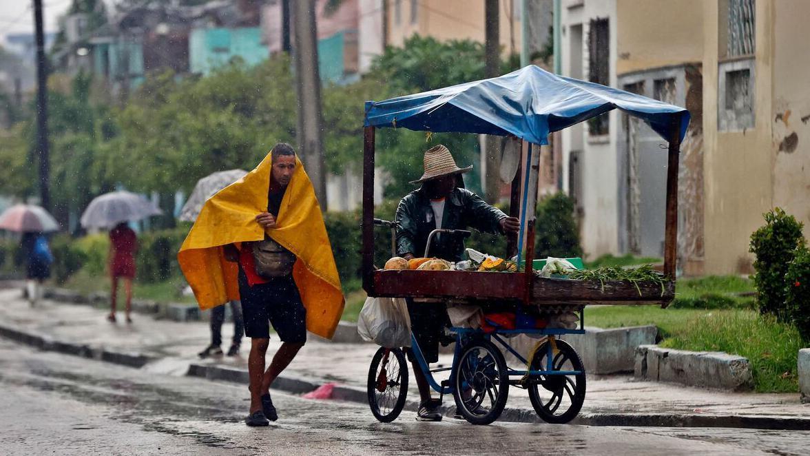 Najmanje 20 mrtvih na Haitiju, uragan Melisa stigao do Kube 3 Dvojica muškaraca se sklanjaju od poplave, jedan je pokriven žutom ceradom, dok drugi vozi kolica sa hranom