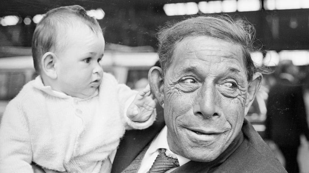Beba u belom kardiganu nežno dodiruje uvo čoveka koji je drži na crnobeloj fotografiji. Čovek, obučen u sako i kravatu, skreće pogled sa deteta. Oboje su među stanovnicima ostrva Tristan da Kunja koji su nedavno stigli u Veliku Britaniju nakon vulkanske erupcije 1961. godine.
