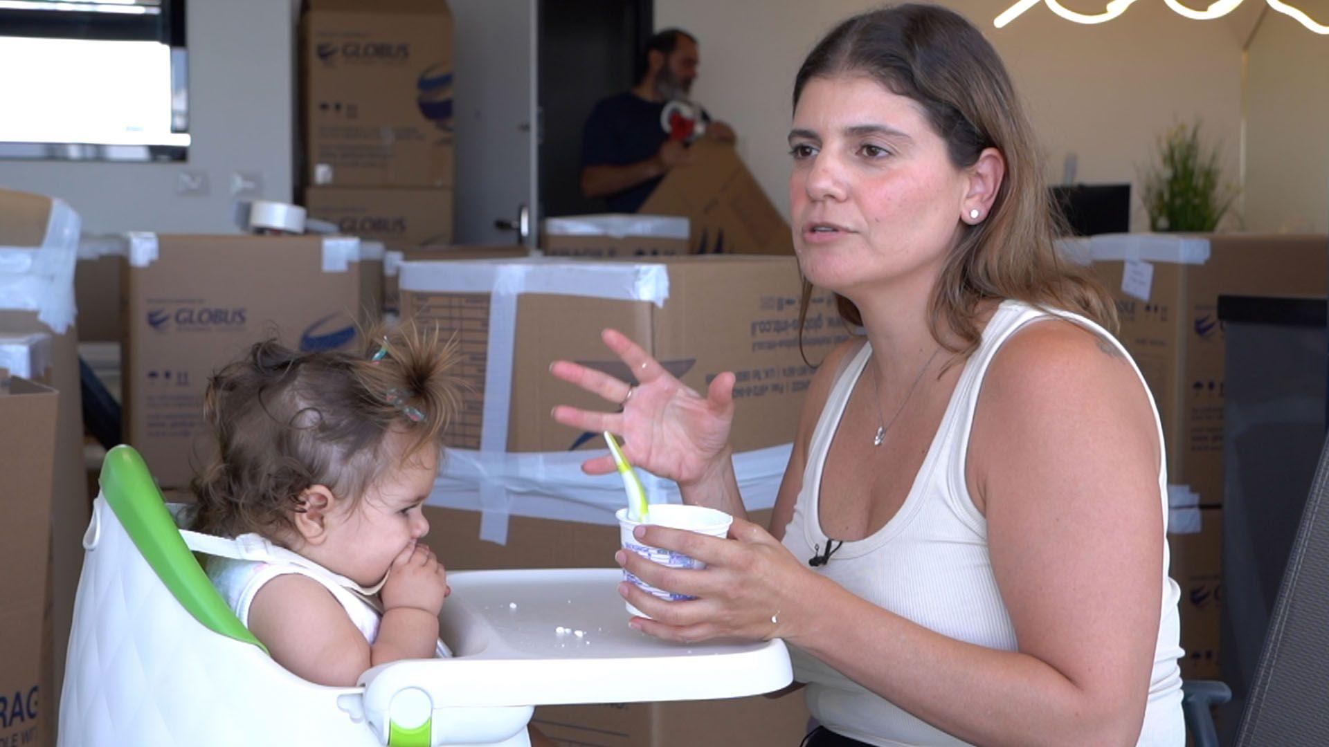 Žena sa smeđom kosom do ramena, obučena u kremasti potplet, hrani malo dete koje sedi u stolici za hranjenje. U pozadini je mnogo kartonskih kutija