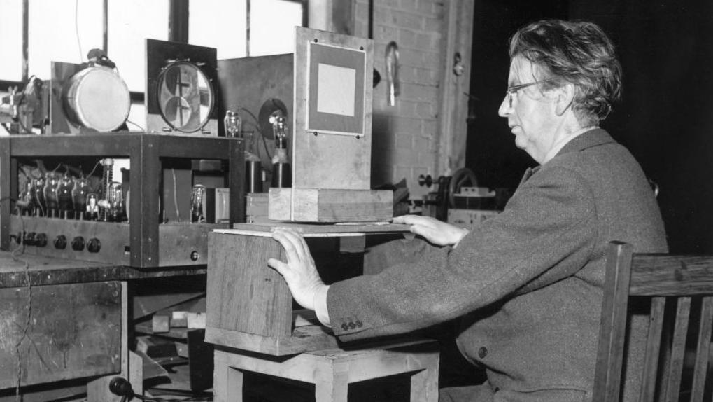 Džon Logi Berd na crno beloj fotografiji sedi sa svojim izumima, okrenut iz profila i radi 