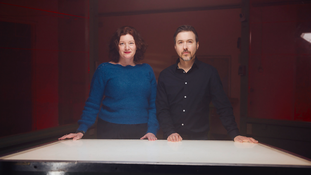 Turi King, u plavoj bluzi dugih rukava, i Aleks Kej, u crnoj košulji, stoje za stolom.