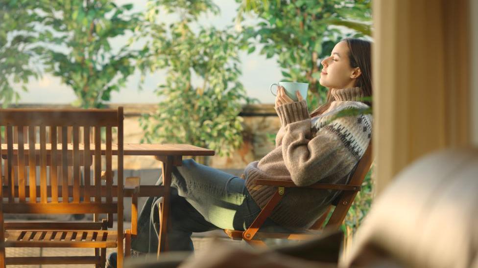 Kako protiv jesenje melanholije 2 Žena u džemperu sedi na terasi ispred kuće, drži piće u šolji i opuštena je sa zadovoljnim izrazom lica.