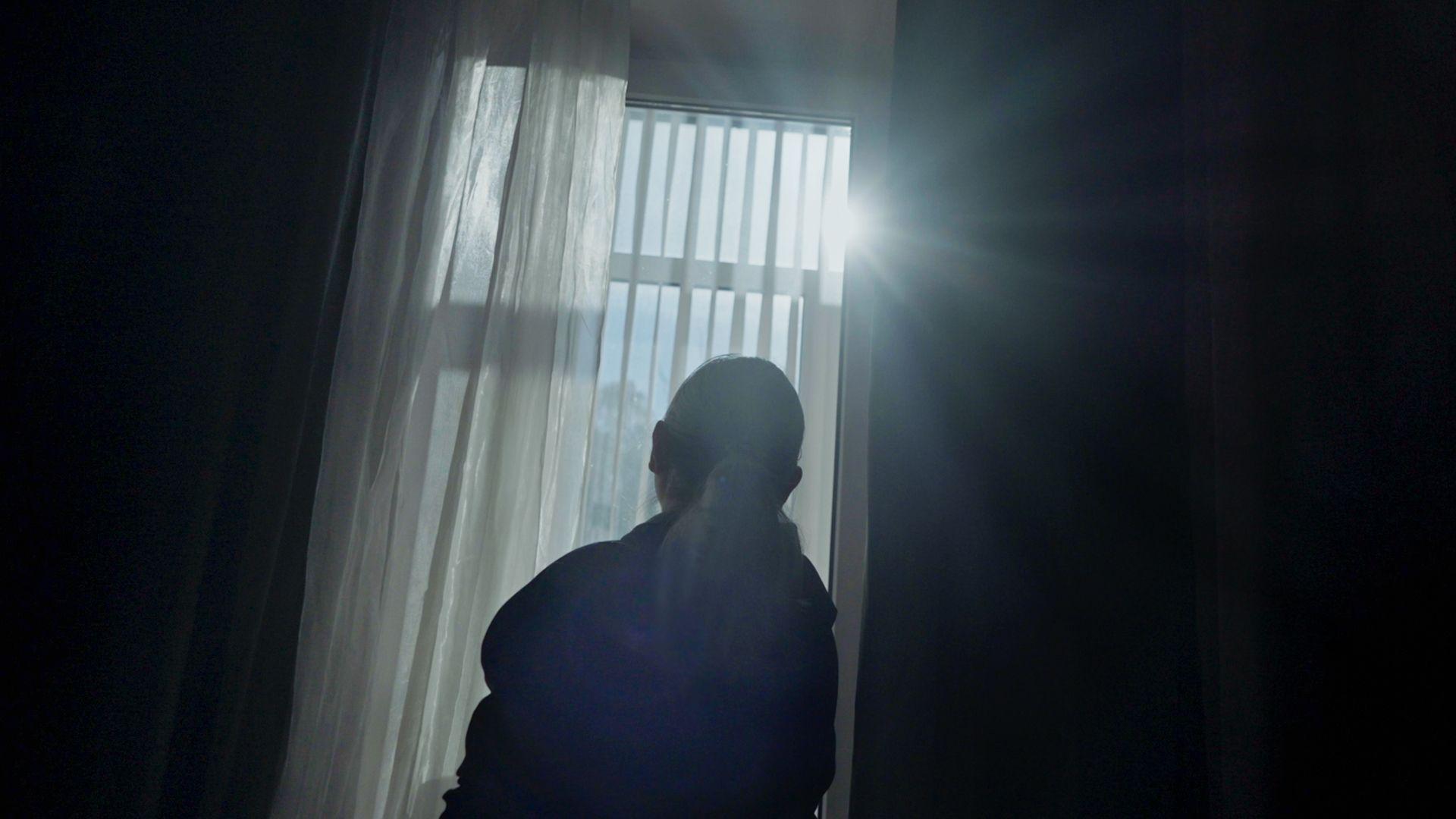 Женщина у зашторенного окна, через которое проглядывает солнце