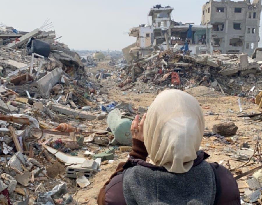 Žena sa maramom na glavi i sivo-smeđem kaputu stoji leđima okrenuta kameri. Ispred nje su ostaci dve uništene betonske zgrade i velike gomile šuta i zemlje