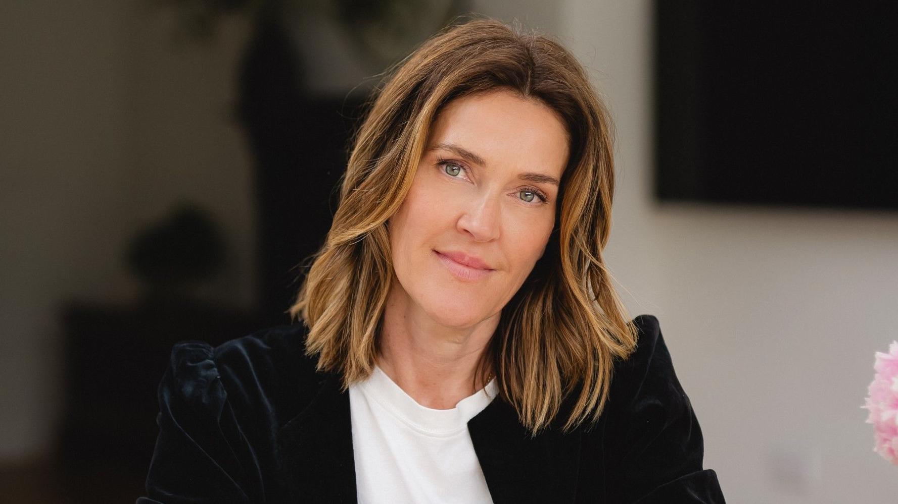 Portret Eme Bardvell, koja ima talasastu kosu sa plavim pramenovima, dugu malo ispod ramena. Smeši se, nosi beli majicu i crni somotski sako.