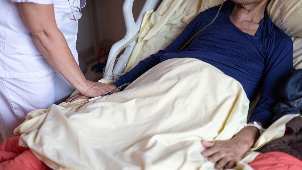 pacijent leži u bolničkom krevetu, medicinska radnica ga drži za ruku 