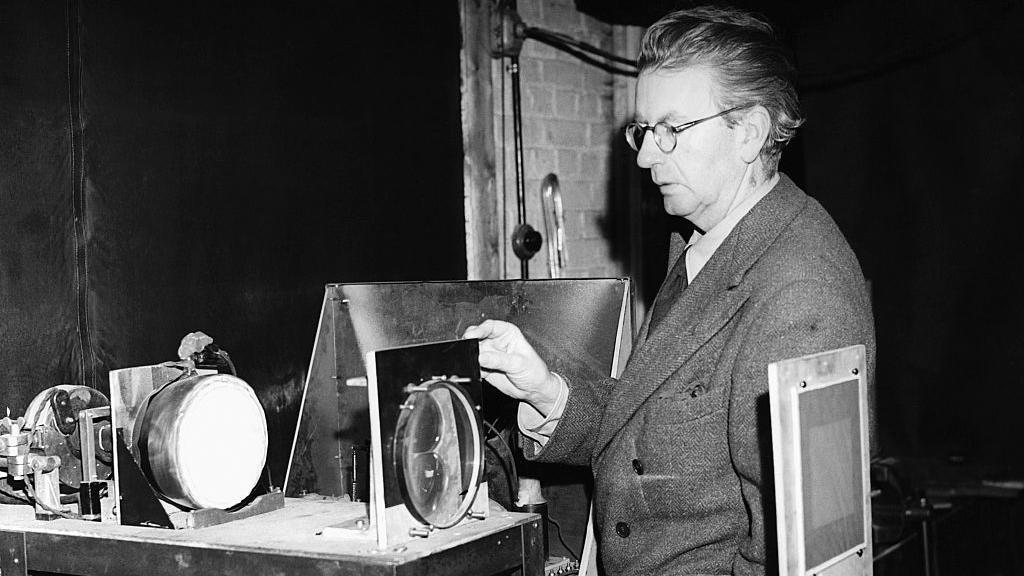 Džon Logi Berd nosi naočale, obučn je u odelo i pokazuje svoje izume na crno-beloj fotografiji