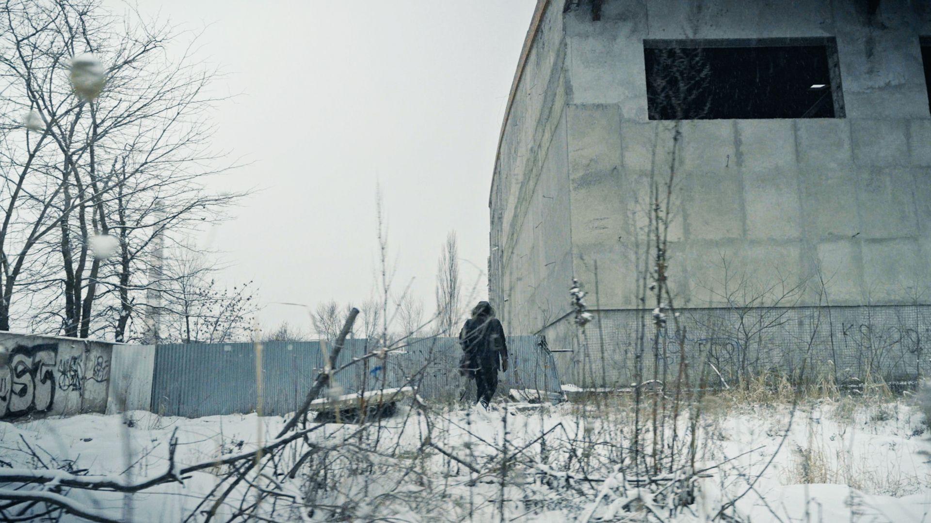 Мужчина с сумкой идет по промзоне зимой мимо здания с темным незастекленным окном в серой бетонной стене
