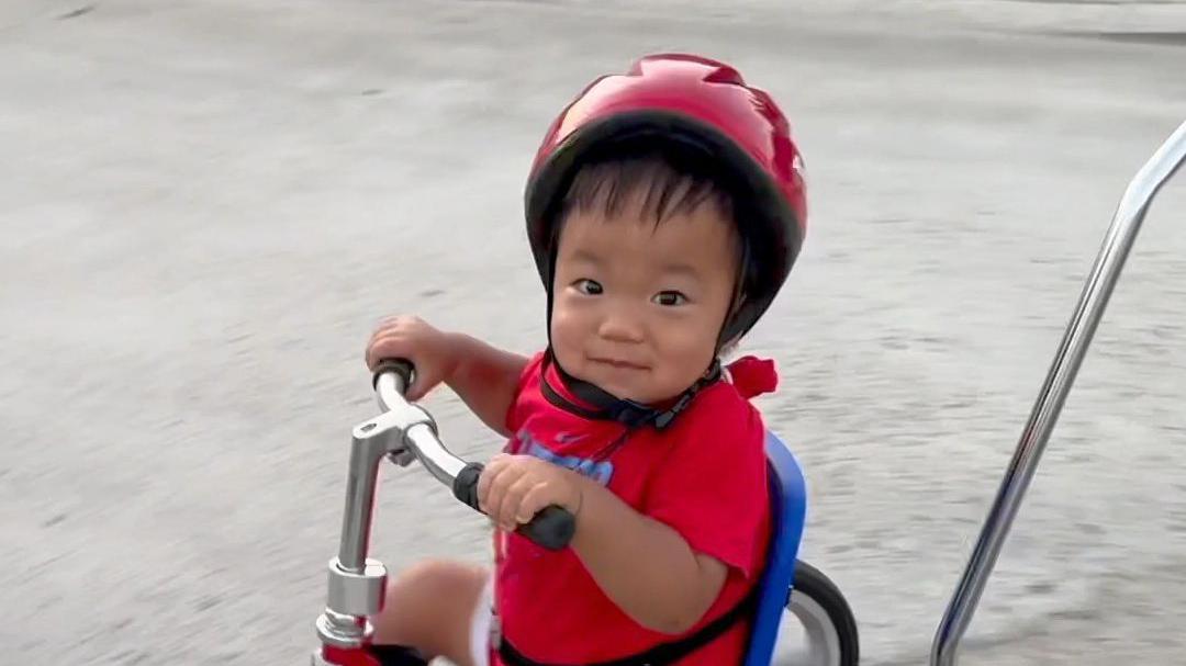 Oliver Ču vozi crven tricikl. Nosi crvenu kacigu, crvenu majicu i crveno-beli šorc i gleda u kameru.