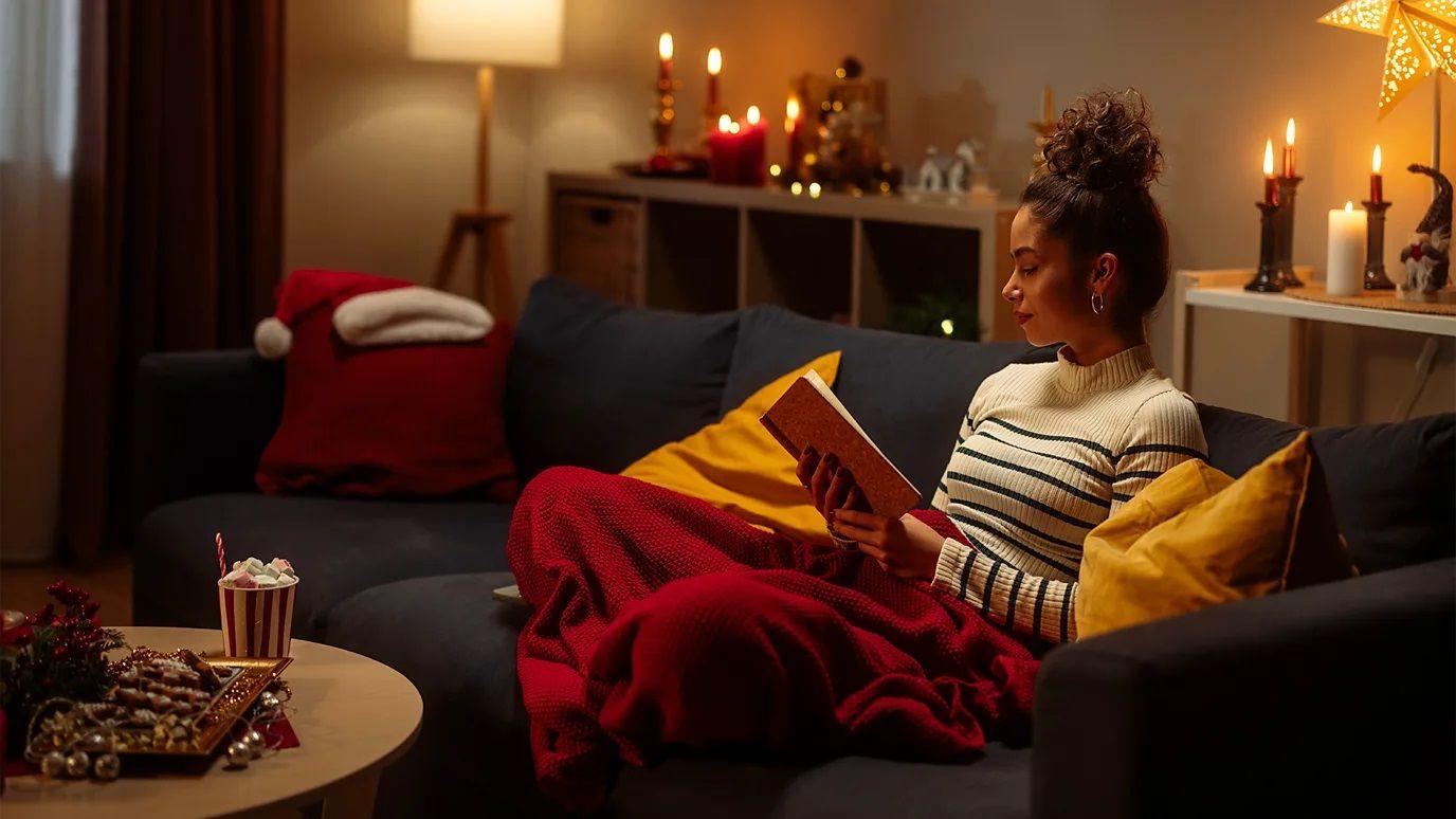 devojka sedi na krevetu prekrivena ćebetom i čita knjigu