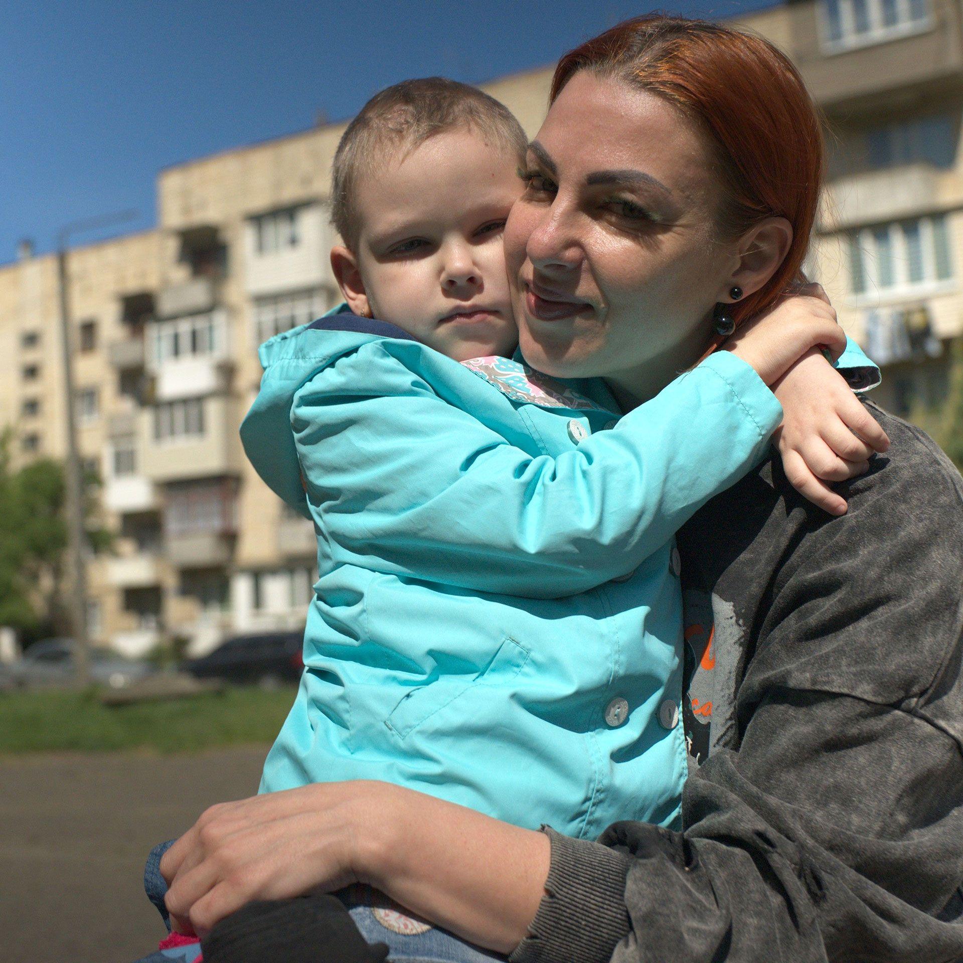 Olena ima ofarbanu crvenu kosu, vezanu pozadi, i nosi sivu majicu. Grli Viktoriju, koja nosi tirkiznu jaknu i ima kratko ošišanu kosu. Nalaze se napolju, a iza njih je stambena zgrada.