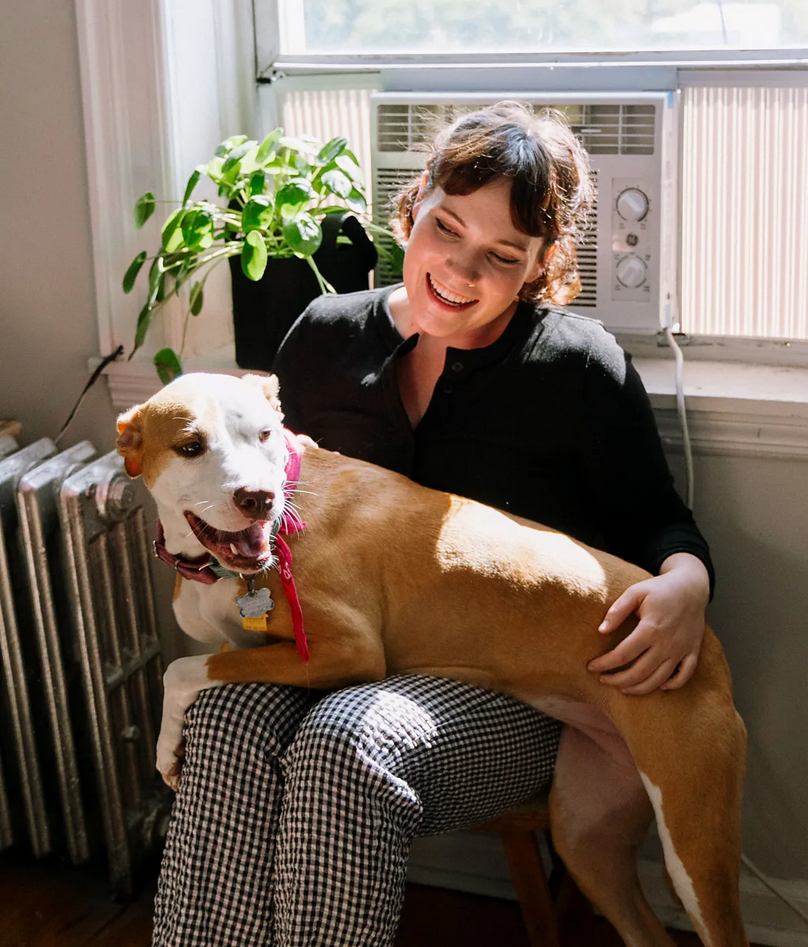 Smiruje li muzika životinje 2 Devojka tamnije vezane kose sedi pored prozora i u krilu drži psa, iza nje su zelena biljka i radijator