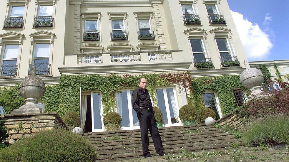 Muškarac obučen u crno stoji na stepenicama ispred luksuzne kuće