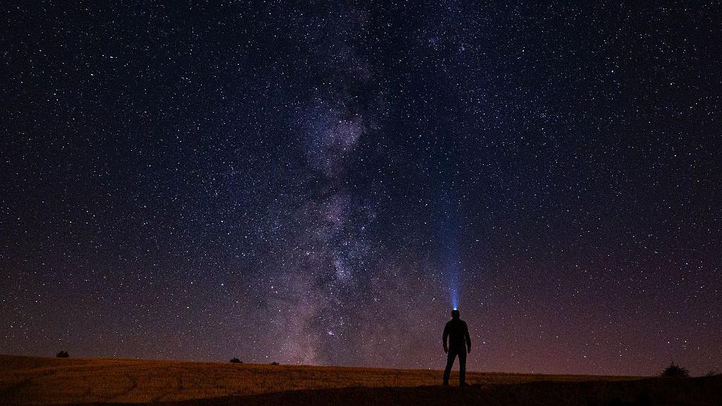 Mala silueta osobe sa lampom na čelu, koja gleda u ogromno noćno nebo ispunjeno bezbrojnim zvezdama, sa galaksijom Mlečni put koja se uzdiže u sredini