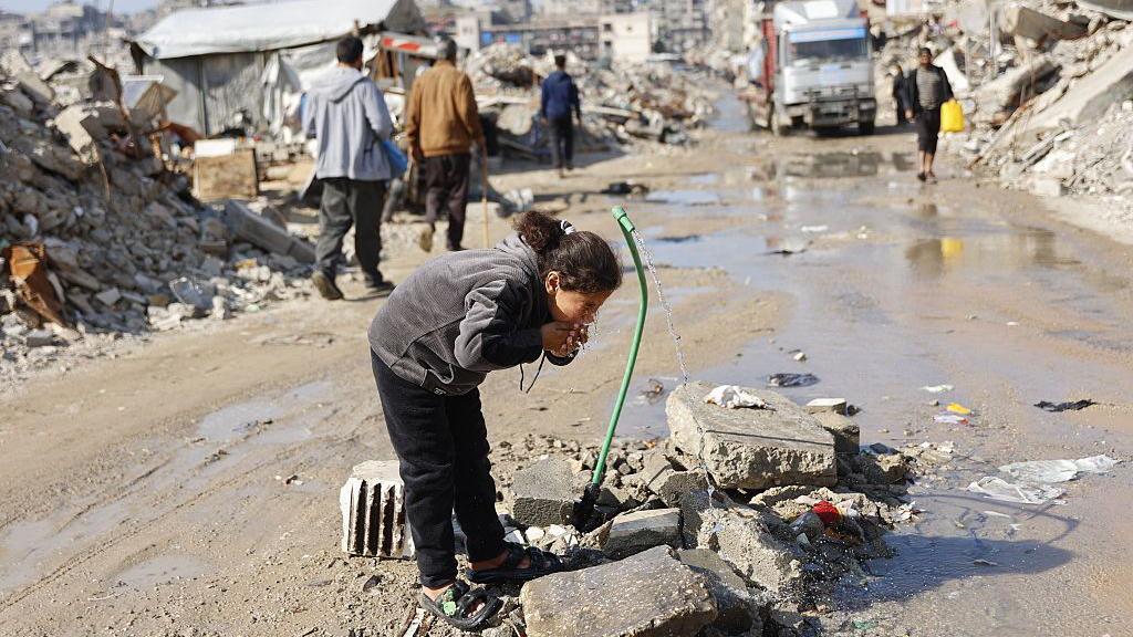 Sve kontroverze Trampovog 'Odbora za mir': Šta je, koga je pozvao i ko je prihvatio 5 A young girl wearing a grey top and black trousers drinks from a green hosepipe surrounded by rubble and debris of homes and businesses in Gaza, with a lorry and pedestrians in the background.