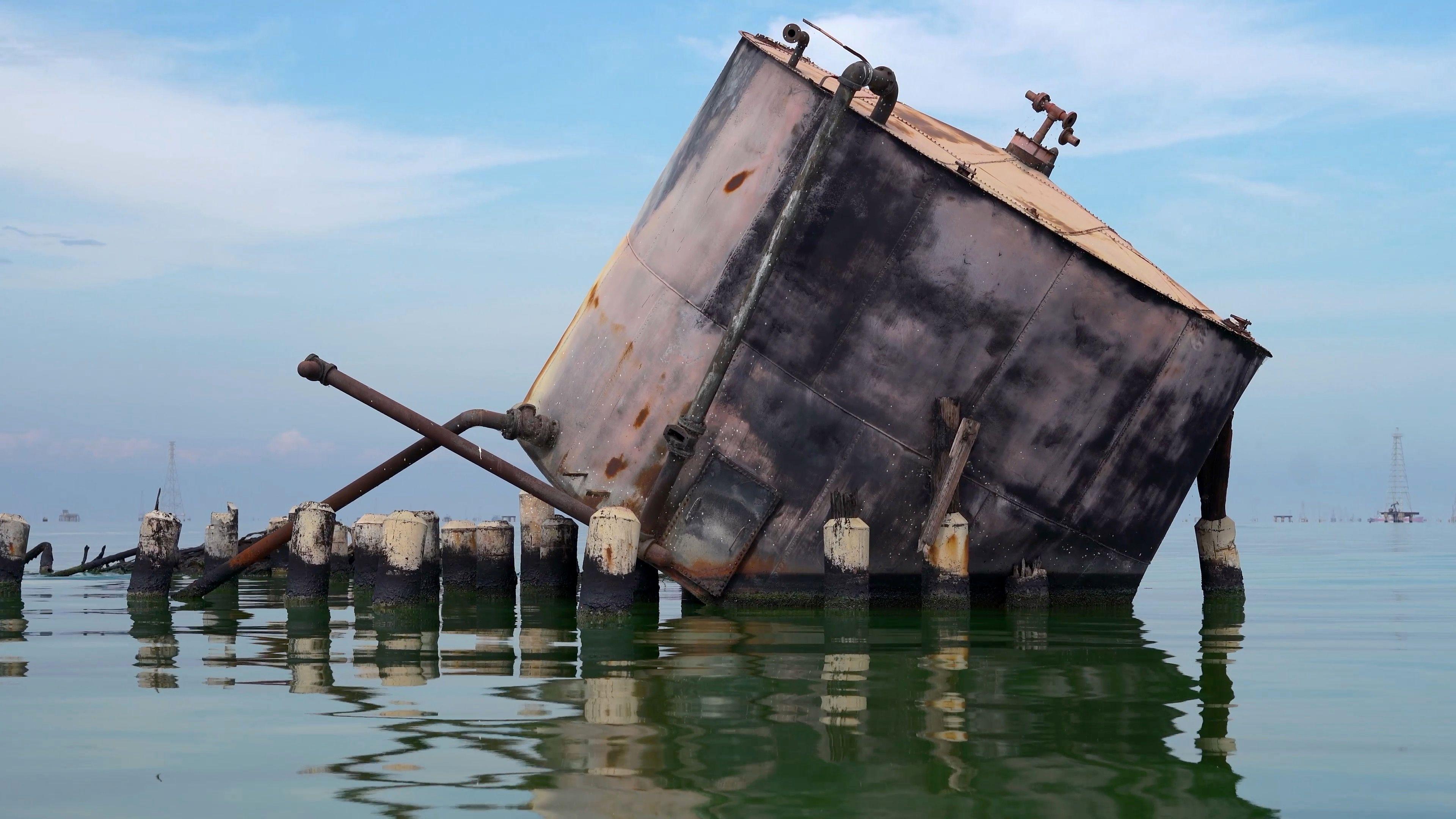 Ko se u Venecueli uzda u Trampa da će doneti oporavak naftne industrije 4 Napušteni, veliki, okrugli metalni rezervoar sa isturenim cevima nalazi se na nivou vode. Nagnut je tako da je deo osnove i bočne strane sa desne strane potopljen. Kratki, debeli stubovi sa prljavo belim vrhovima vire iz vode ispred rezervoara.