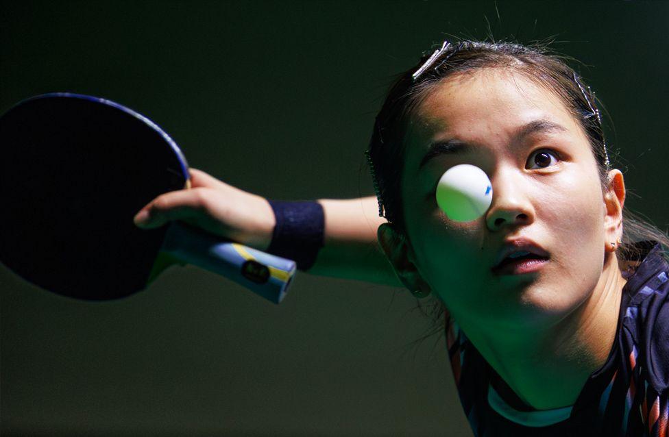 Krupni plan stonoteniserke koja se sprema da reketom udari lopticu, ruka joj je ispružena i na njoj se vidi narukvica. Loptica se vidi u letu iznad desnog oka igrača.