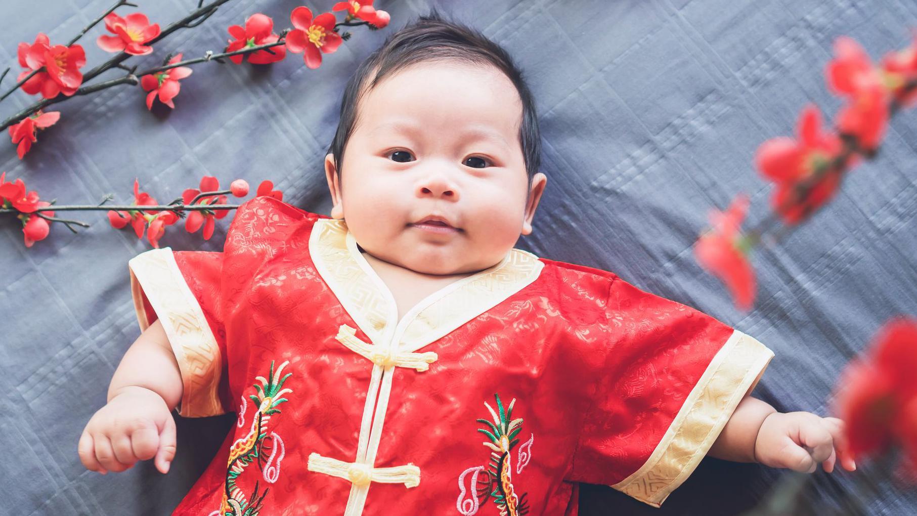 A baby lying down on a patterned grey cloth while dressed in a red traditional Chinese outfit with gold linings. Some red flowers surround him. 