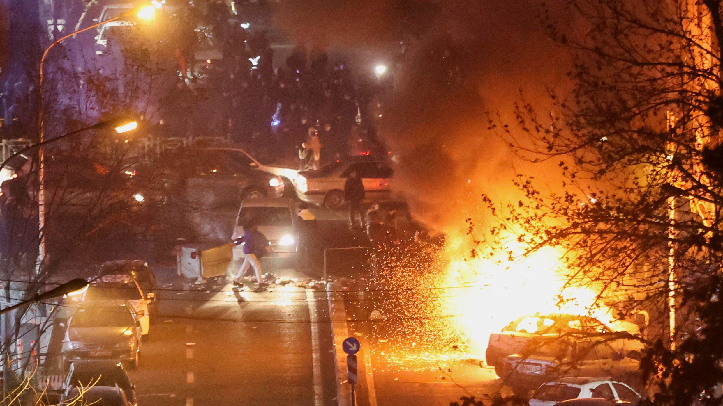 Vatra gori tamo gde su parkirani automobili u Teheranu 8. januara 2026. Ljudi su okupljeni u blizini. Drvo je u prvom planu.